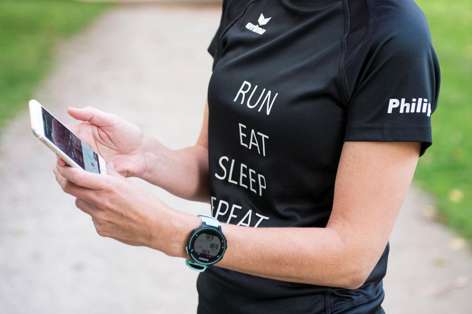 Frau mit kurzer blonder Frisur schaut auf ihr Smartphone, trägt ein Sport-T-Shirt mit Schriftzug und eine Sportuhr am Handgelenk.