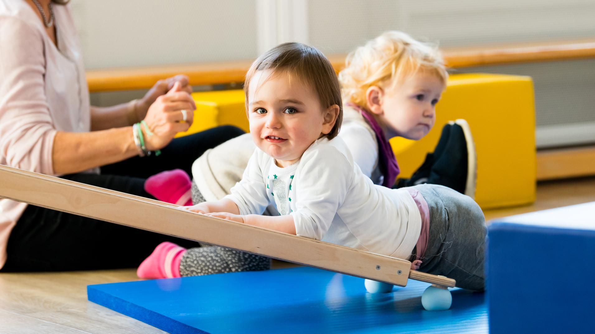 Zwei Kleinkinder spielen auf einer blauen Matte, während eine betreuende Person im Hintergrund sitzt.